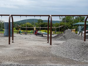 A wide view of a newly installed playground shows multiple play features set against a backdrop of green trees and rolling hills. Brown metal swing frames stretch across the foreground, with chains hanging down but no swing seats attached. The playground base consists of a broad expanse of light gray crushed stone or compacted gravel, covering the entire play area beneath and around the equipment. The surface appears freshly spread, level and free of vegetation, providing a uniform base for the playground. Play elements include accessible seating spinners, climbing structures, balance features, ramps and shaded gathering areas. The gravel surface extends throughout the playground footprint.