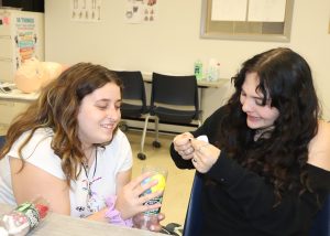 Two Capital Region BOCES EMT students work together at a classroom table while building a model lung from a plastic bottle, balloons and other materials during a lesson on the respiratory system. A CPR training mannequin is visible in the background.
