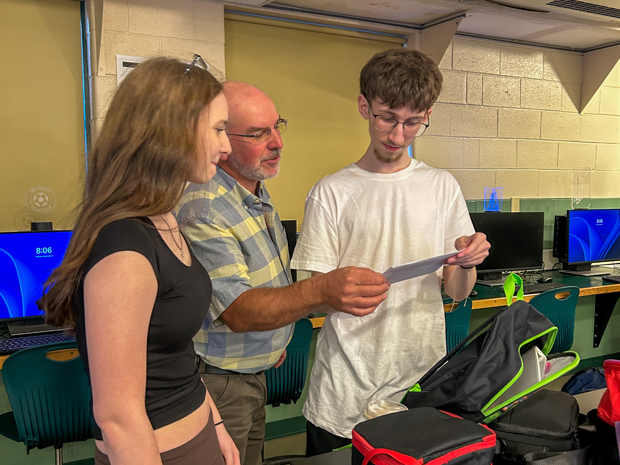 A teacher and two students stand in a computer lab reviewing a sheet of paper together, with desktop computers and classroom seating visible in the background