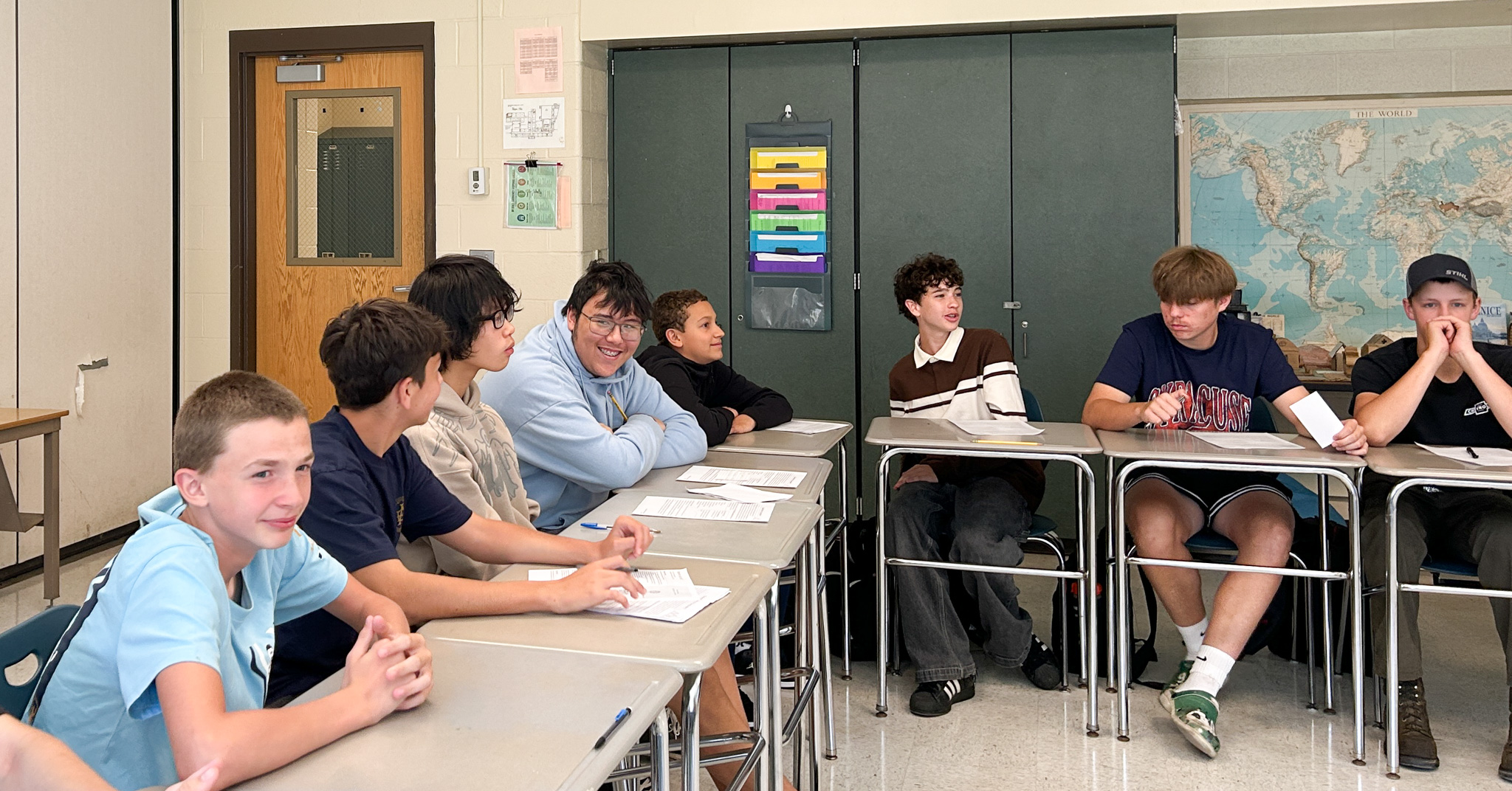 Students sit at desks arranged in a U-shape during a classroom discussion, with papers on their desks and a world map on the wall behind them.