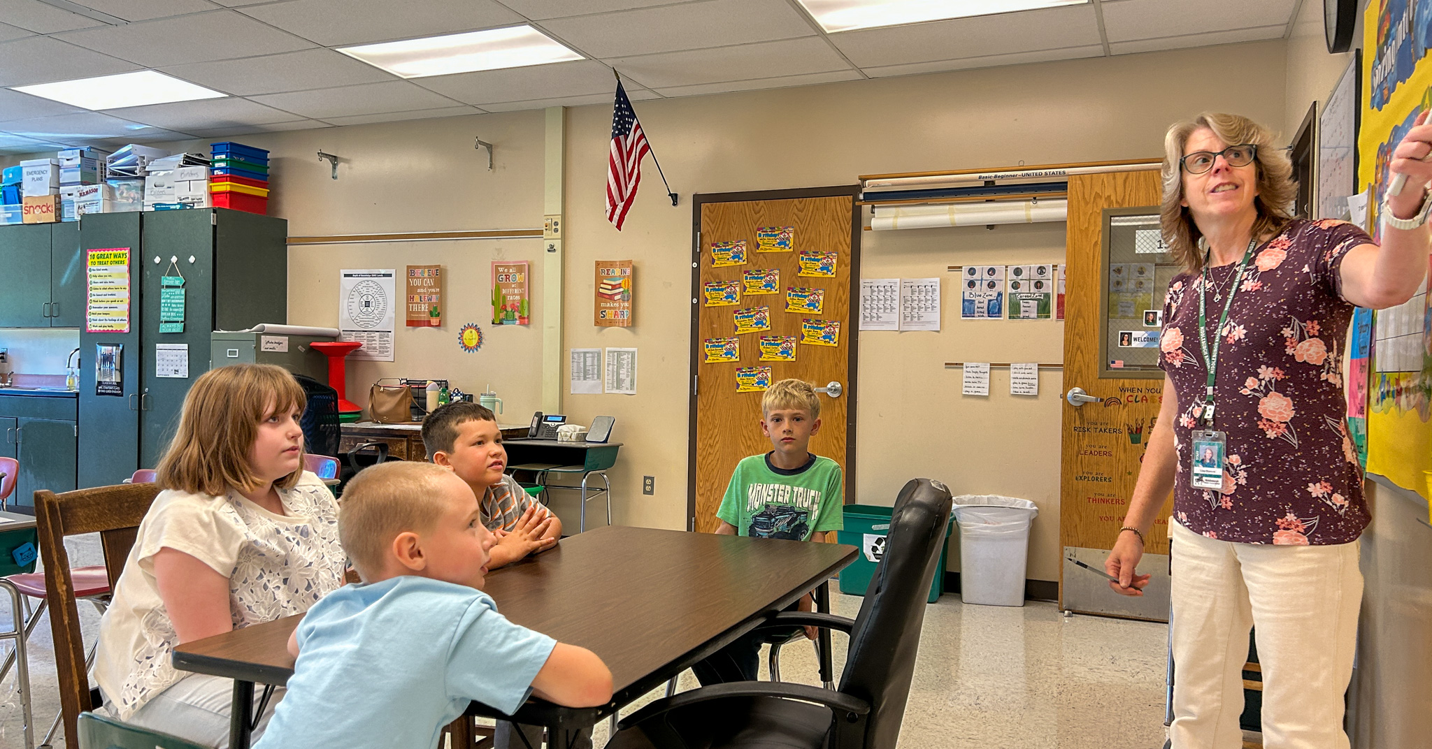 An educator points to a classroom display while several elementary students sit around a table and look toward the front of the room.