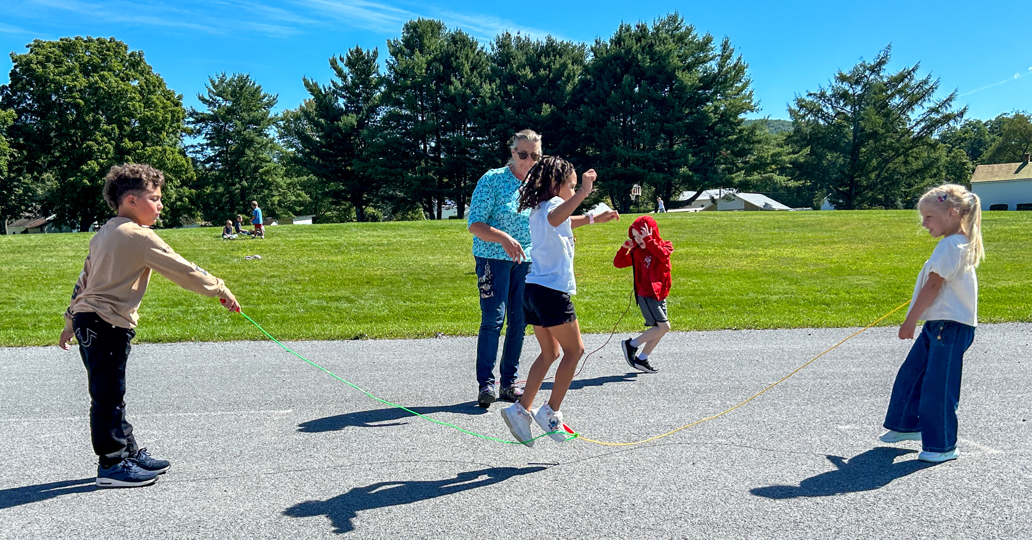 Children participate in a jump rope activity outdoors while an adult supervises on a paved area beside a grassy field and trees