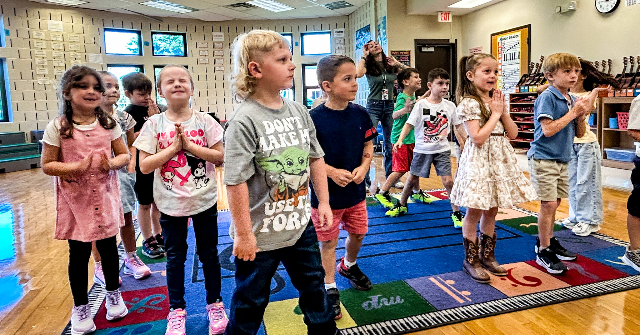 Young students stand on a colorful classroom rug and take part in a music and movement activity while an instructor leads from the back of the room.