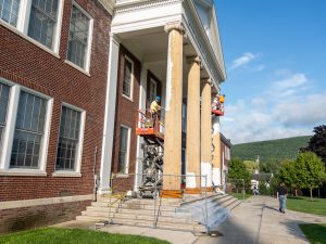 Painters wearing hard hats stand on lifts as they apply paint to bare wood of columns.