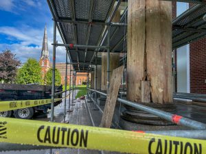 Scaffolding in front of bare wood columns after paint removal. Decay is visible in some parts of the wood. Caution tape surrounds project.
