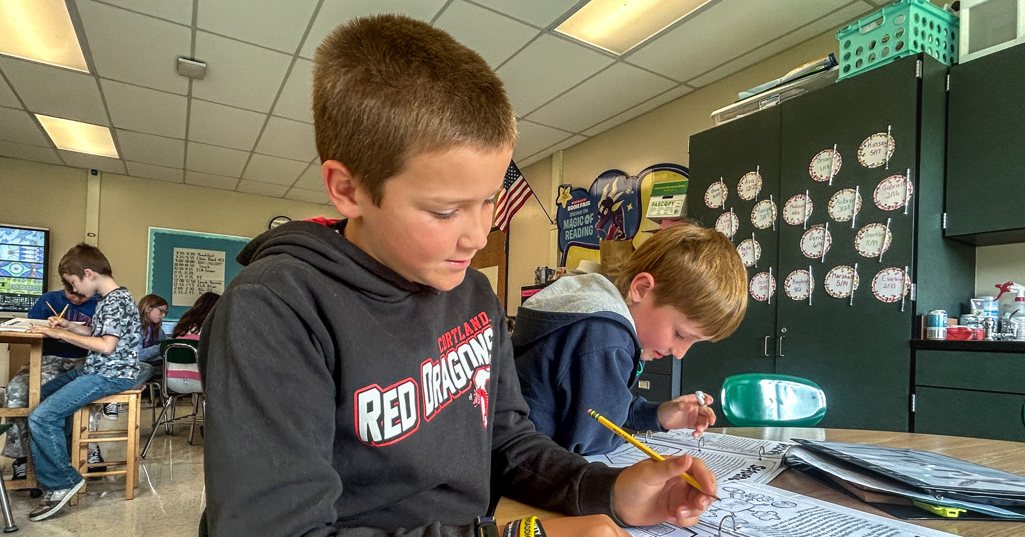 Two elementary students work with pencils and binders at a table. Additional students work in the background of the classroom.
