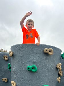 A child in a bright orange T-shirt stands at the top of a gray playground climbing structure with green and tan handholds, raising one hand overhead. The photo is taken from below against a cloudy gray sky.