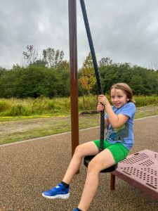 A child sits on zip line at a playground, holding the rope with both hands. Trees and grassy fields are visible in the background beneath an overcast sky.