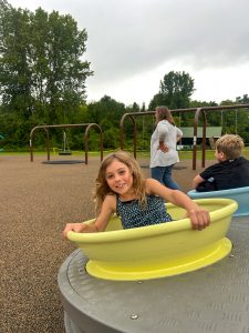 A child sits in a light green spinning playground bowl on a large round play feature. A playground with swings, trees and other visitors appears in the background under cloudy skies.