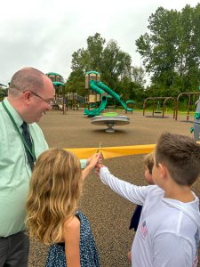 Middleburgh superintendent Mark Place and three children participate in a ribbon-cutting ceremony at a playground, holding ceremonial scissors to cut a yellow ribbon. 