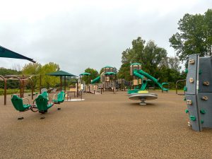 Wide view of an accessible playground with a poured-in-place rubber safety surface in shades of tan and brown. The playground features a large multi-level play structure with ramps, elevated platforms, tunnel slides, straight slides and a rope climbing bridge beneath green shade roofs. In the foreground are molded bucket-style spinner seats mounted on a rotating play element and a large circular spinning platform with a bowl seat. A freestanding gray climbing wall with green and tan handholds stands at right, while swing frames, shade canopies and additional play features are visible throughout the play area. Trees and an overcast sky form the backdrop.