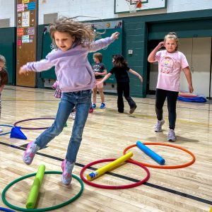 Elementary students participate in a physical education activity, moving through an obstacle course made of hula hoops and foam noodles in the school gymnasium.