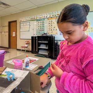 A second grade student works on a cardboard community project, carefully placing materials inside a handmade cardboard building during a social studies lesson about government and community planning.