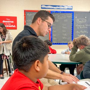 A student and teacher work together at a classroom table. Other students are in the background.