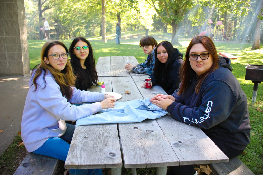 Five members of the Class of 2027 sit together at a picnic table in a park. A pair of jeans is on the table.